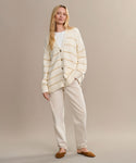 A woman with long blonde hair poses casually against a beige backdrop, wearing light beige pants, a cream and beige striped cardigan over a white top, and Italian-made Clara Mule slippers in cognac.