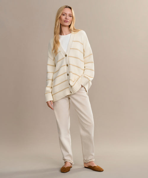 A woman with long blonde hair poses casually against a beige backdrop, wearing light beige pants, a cream and beige striped cardigan over a white top, and Italian-made Clara Mule slippers in cognac.