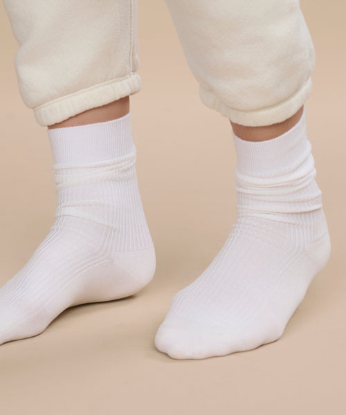 A person stands on a beige surface, wearing off-white sweatpants with rolled cuffs that reveal their feet in Classic Socks, ivory color.