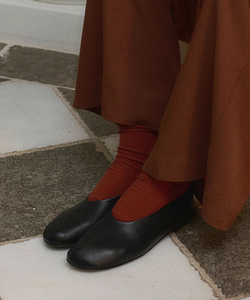 A person wearing wide brown pants, Classic Sock in rust lyocell blend, and black ballet flats stands on a tiled floor with a stone and white pattern.