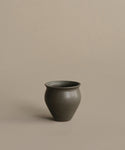 The Classic Vessel is a handmade dark gray vase featuring a rounded body and slightly flared rim, displayed on a plain beige surface with a matching background.