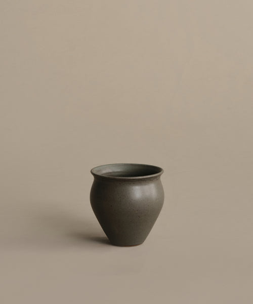 The Classic Vessel is a handmade dark gray vase featuring a rounded body and slightly flared rim, displayed on a plain beige surface with a matching background.