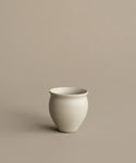 The Classic Vessel, a simple off-white stoneware piece with a rounded shape, sits on a plain beige surface against a matching beige background.