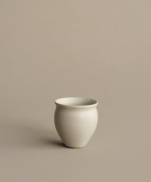 The Classic Vessel, a simple off-white stoneware piece with a rounded shape, sits on a plain beige surface against a matching beige background.