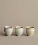 Three Classic Vessel stoneware cups, simple and round with an off-white finish, are arranged in a row on a beige background. Slight variations in shape and rim emphasize their handcrafted quality.