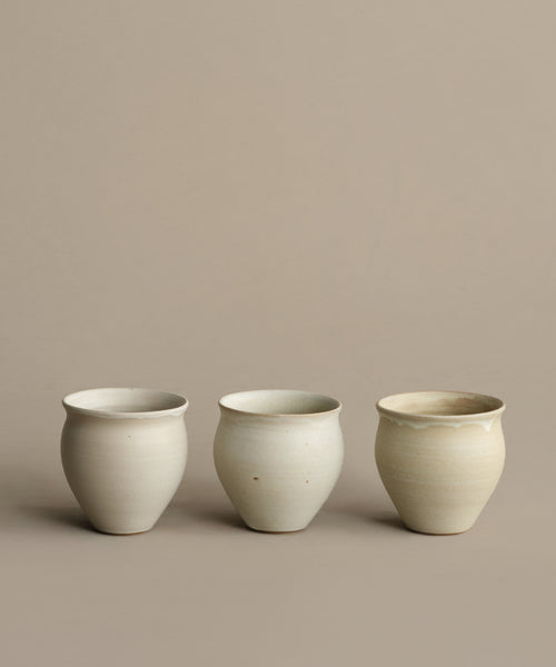 Three Classic Vessel stoneware cups, simple and round with an off-white finish, are arranged in a row on a beige background. Slight variations in shape and rim emphasize their handcrafted quality.