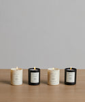 The Votive Candle Set features four hand-poured candles—two white and two black—in glass jars with white scent-detail labels, arranged on a wooden surface against a gray background for a stylish display.