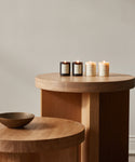 Two round wooden tables of different heights display a ceramic bowl on the lower table and the Votive Candle Set, featuring four hand-poured glass candles, on the taller table, all set against a neutral wall.