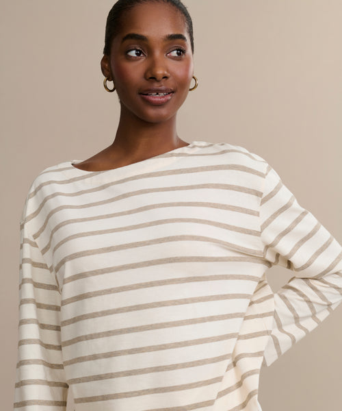 A woman with dark skin and hoop earrings smiles softly. She wears the Claude Long-Sleeve Tee, made of 100% cotton, featuring light beige nautical stripes on a neutral background.
