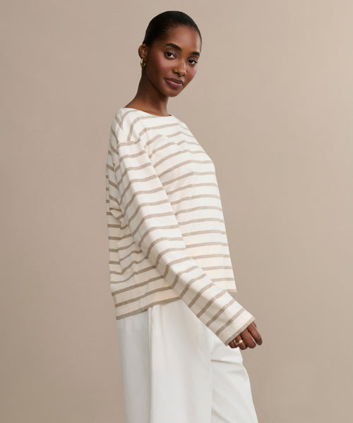 A woman stands against a beige background, smiling gently at the camera with her hair pulled back. She wears the Claude Long-Sleeve Tee—100% cotton with cream and tan nautical stripes—and white pants.