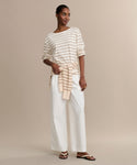 A woman stands against a beige background wearing the Claude Long-Sleeve Tee in cream and tan stripes, paired with white wide-leg pants, sandals, and a matching striped sweater tied at her waist.