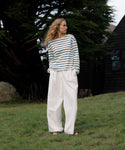 Woman wearing a striped shirt and white pants standing in a grassy area with trees and a building in the background.
