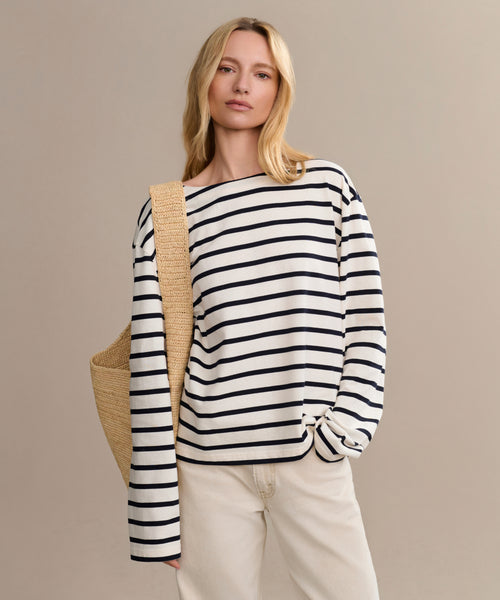 A blonde woman wears the Claude Long-Sleeve Tee with nautical stripes and 100% cotton, paired with light beige pants and a woven straw bag, standing against a plain beige background.