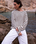 A person with short dark hair wearing the Claude Long-Sleeve Tee and white pants sits on rocks by the water, looking to the side with a rocky shoreline in the background.