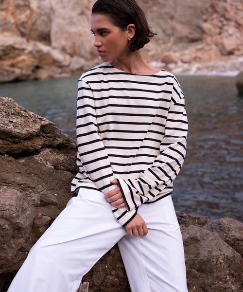 A person with short dark hair wearing the Claude Long-Sleeve Tee and white pants sits on rocks by the water, looking to the side with a rocky shoreline in the background.
