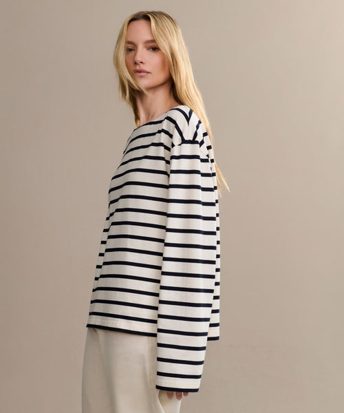 A woman with long blonde hair wears the Claude Long-Sleeve Tee—an oversized, 100% cotton top with black nautical stripes—and loose beige pants, standing against a plain beige background.