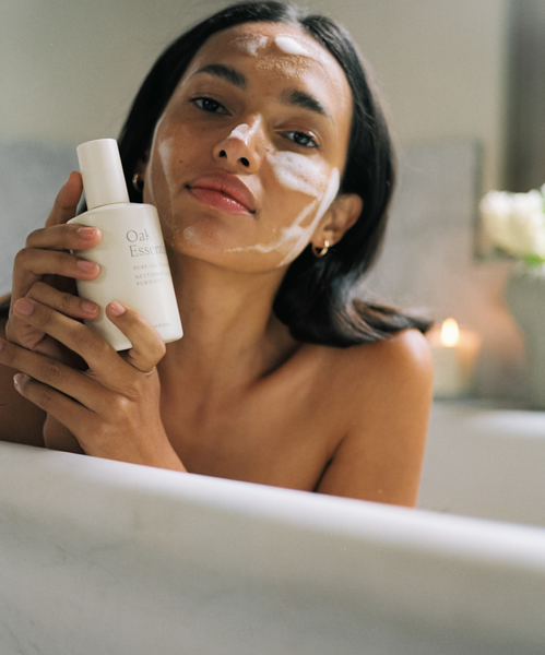 A woman relaxes in a bathtub, holding the Cleanse and Polish Duo. She has cream on her face for a radiant glow, with a candle and flowers softly blurred in the background.