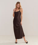 A woman with side-parted blonde hair stands against a plain background in the Cleo Slip Dress, a sleeveless dark brown, ankle-length, bias cut dress. She pairs it with black flats and gently holds the side of her dress while looking down.