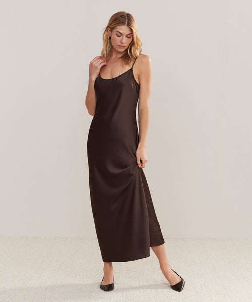 A woman with side-parted blonde hair stands against a plain background in the Cleo Slip Dress, a sleeveless dark brown, ankle-length, bias cut dress. She pairs it with black flats and gently holds the side of her dress while looking down.