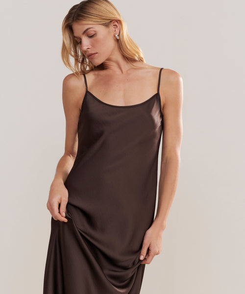 A woman with blonde hair wears the Cleo Slip Dress, a sleeveless dark brown satin dress with thin straps. She stands against a plain light background, looking down and gently holding part of the dress.