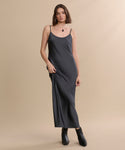 A woman with short brown hair wears the Cleo Slip Dress, a sleeveless dark gray satin style, paired with black ankle boots, standing relaxed against a beige background.