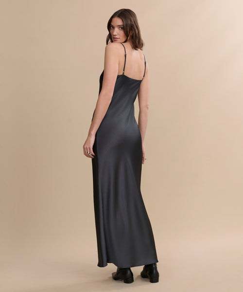 A woman stands facing away, looking over her shoulder. She wears the Cleo Slip Dress, a long black slip with thin straps and a bias cut, paired with black boots against a beige background.