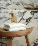 A bottle of Cloud Foaming Bath Oil with a wooden cap rests on white towels atop a wooden stool, set against a marble-patterned bathroom background for a relaxing, spa-like experience.
