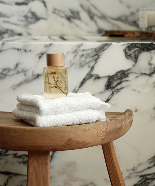 A bottle of Cloud Foaming Bath Oil with a wooden cap rests on white towels atop a wooden stool, set against a marble-patterned bathroom background for a relaxing, spa-like experience.