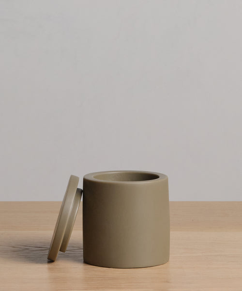 The Coast Canister, a cylindrical matte taupe handcasted resin container with matching lid, rests on light wood in front of a plain light grey background as part of the Coast Collection.