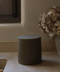 A matte dark Coast Canister from the Coast Collection, made of handcasted resin with a fitted lid, sits on a beige kitchen countertop beside a vase of dried hydrangeas near a window.