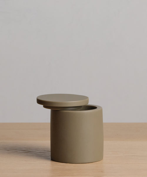A small, minimalist Coast Canister from the Coast Collection, made of hand-cast resin, rests with a slightly open round lid on a light wooden surface against a plain light gray background.