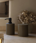 The Coast Canister and matte brown soap dispenser from the Coast Collection rest on a beige stone countertop, accompanied by dried hydrangeas for a serene, neutral-toned bathroom look.