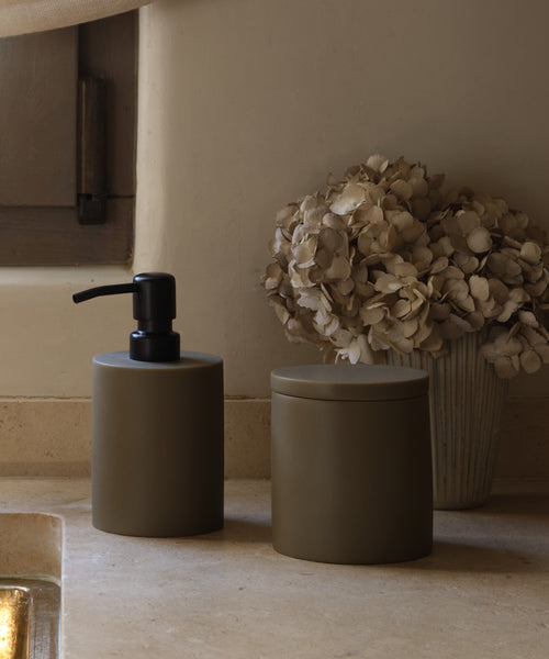 The Coast Canister and matte brown soap dispenser from the Coast Collection rest on a beige stone countertop, accompanied by dried hydrangeas for a serene, neutral-toned bathroom look.