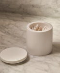 A white, cylindrical Coast Canister rests on a marble countertop, holding cotton swabs. This elegant home accessory comes with a matching lid placed alongside it.