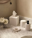 A marble countertop with white home accessories, featuring the Coast Canister, a matching tissue box, pump dispenser, jewelry dish, and a small vase with white daisies.