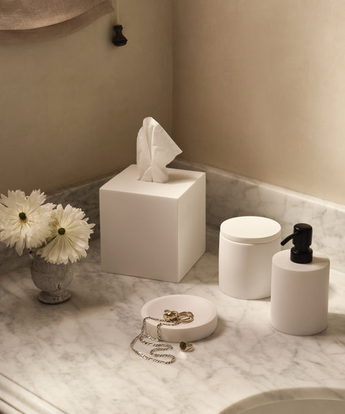A marble countertop with white home accessories, featuring the Coast Canister, a matching tissue box, pump dispenser, jewelry dish, and a small vase with white daisies.