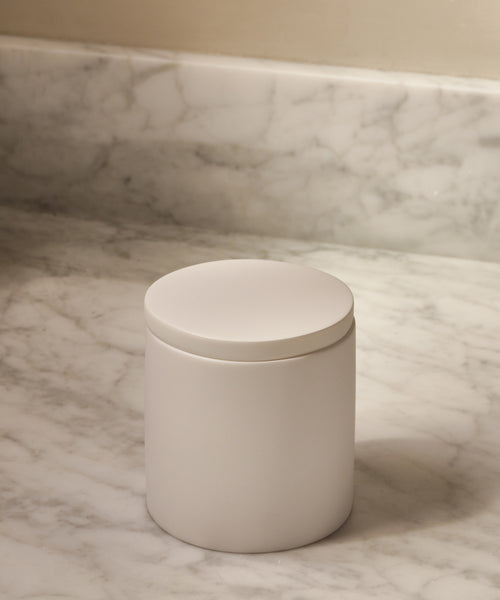 A Coast Canister in handcasted white resin with a round lid rests on a marble countertop and backsplash—an elegant touch to your home accessories collection.