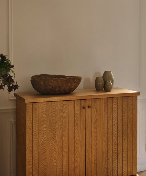 The Coast Hutch, a solid white oak mid-century modern piece with vertical grooves, stands against a white wall. On top, a rustic bowl and two grey vases add style, while dark-leaved greenery is partly visible on the left.