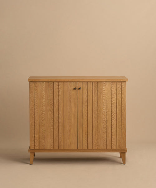 The Coast Hutch is a solid white oak cabinet with vertical slats, two central doors, small black handles, and short angled legs in a mid-century modern hutch design, shown against a beige background.