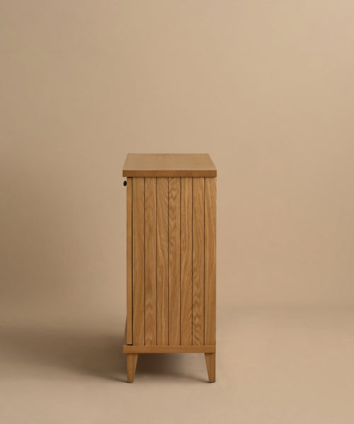 The Coast Hutch is a wooden cabinet with vertical slats, shown from the side against a beige background. Made from solid white oak with a light natural finish, it stands on tapered legs and provides stylish storage for any room.