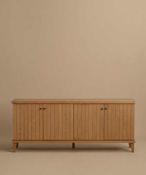 The Coast Sideboard in Warm Oak features vertical panel doors, four small handles, and angled legs, shown against a beige background.