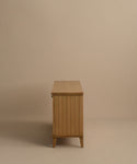 The Coast Sideboard features vertical slats in Warm Oak and short legs, set against a beige background. Its clean, minimalist silhouette is inspired by mid-century modern design.