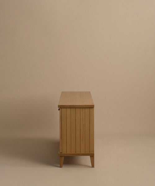 The Coast Sideboard features vertical slats in Warm Oak and short legs, set against a beige background. Its clean, minimalist silhouette is inspired by mid-century modern design.