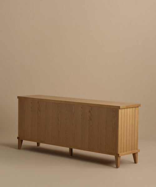 The Coast Sideboard is a Mid-Century Modern wooden piece with a light natural finish, vertical grooves on the sides, and angled legs, beautifully displayed against a beige background.