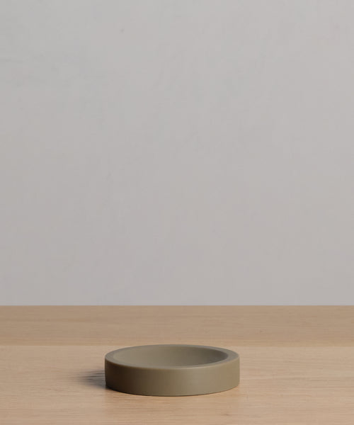 The Coast Soap Dish, a simple round and flat piece made from taupe handcast resin, rests on light wood with a plain light gray backdrop.