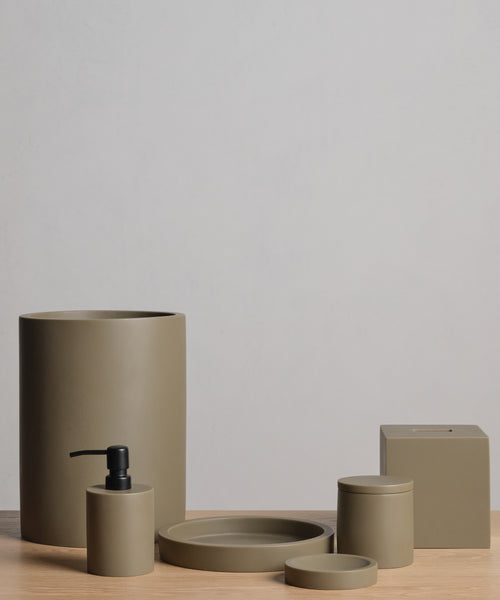 A set of five matte olive green bathroom accessories—handcast in resin, including a waste bin, soap dispenser, tray, lidded jar, and the Coast Soap Dish—displayed on light wood with a neutral background.