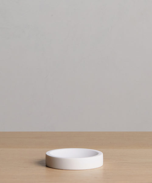 The Coast Soap Dish is a small, round, white resin dish, handcasted in Mexico. It sits on a light wooden surface with a plain, light gray background.