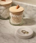 A marble bathroom counter features the Coast Soap Dish, crafted from handcast resin in Mexico, holding three rings. In the background, glass jars with wooden lids store cotton swabs and cotton balls.