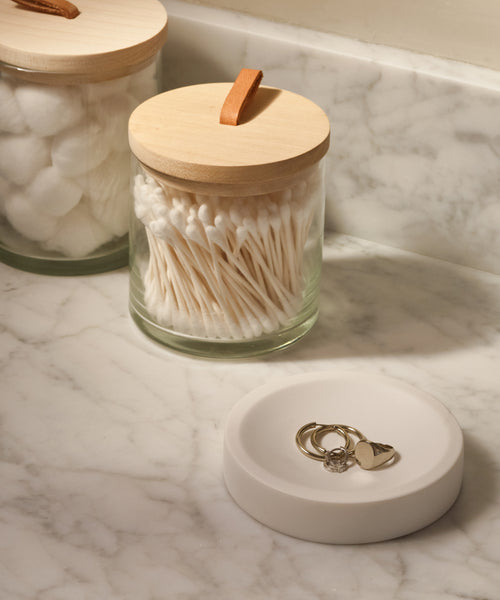 A marble bathroom counter features the Coast Soap Dish, crafted from handcast resin in Mexico, holding three rings. In the background, glass jars with wooden lids store cotton swabs and cotton balls.