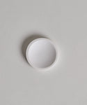 A Coast Soap Dish, small, round, and white, rests on a light gray textured surface with a faint shadow on its left side.